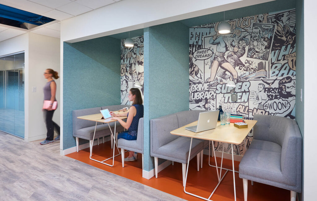 Modern office space with a woman working on a laptop at a booth with comic strip wall art, and another person walking by.