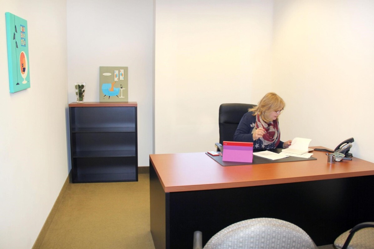 A woman sits at a desk in an office with a phone, papers, a laptop, and a bookshelf. The walls are adorned with two pieces of colorful art.