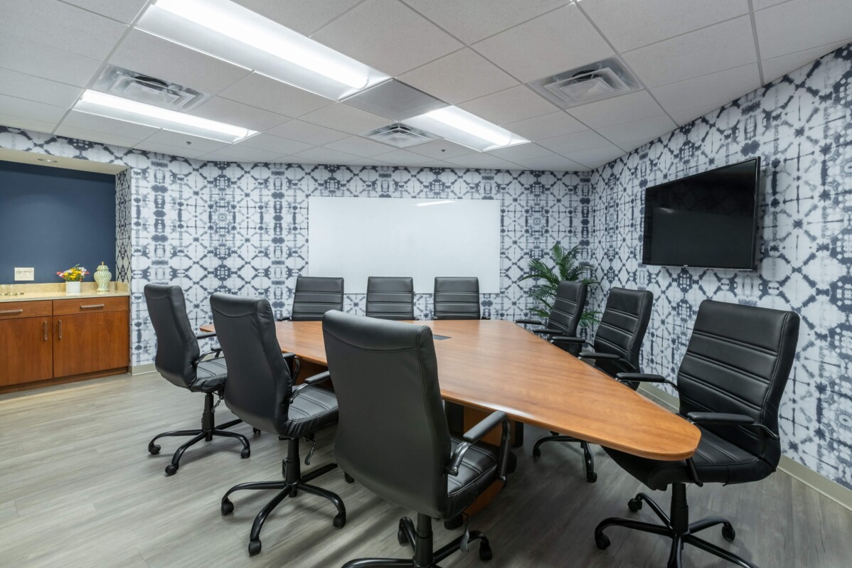 A conference room with a large oval wooden table surrounded by eight black office chairs, a TV mounted on the wall, and a whiteboard. The walls are patterned and a cabinet is in the corner.