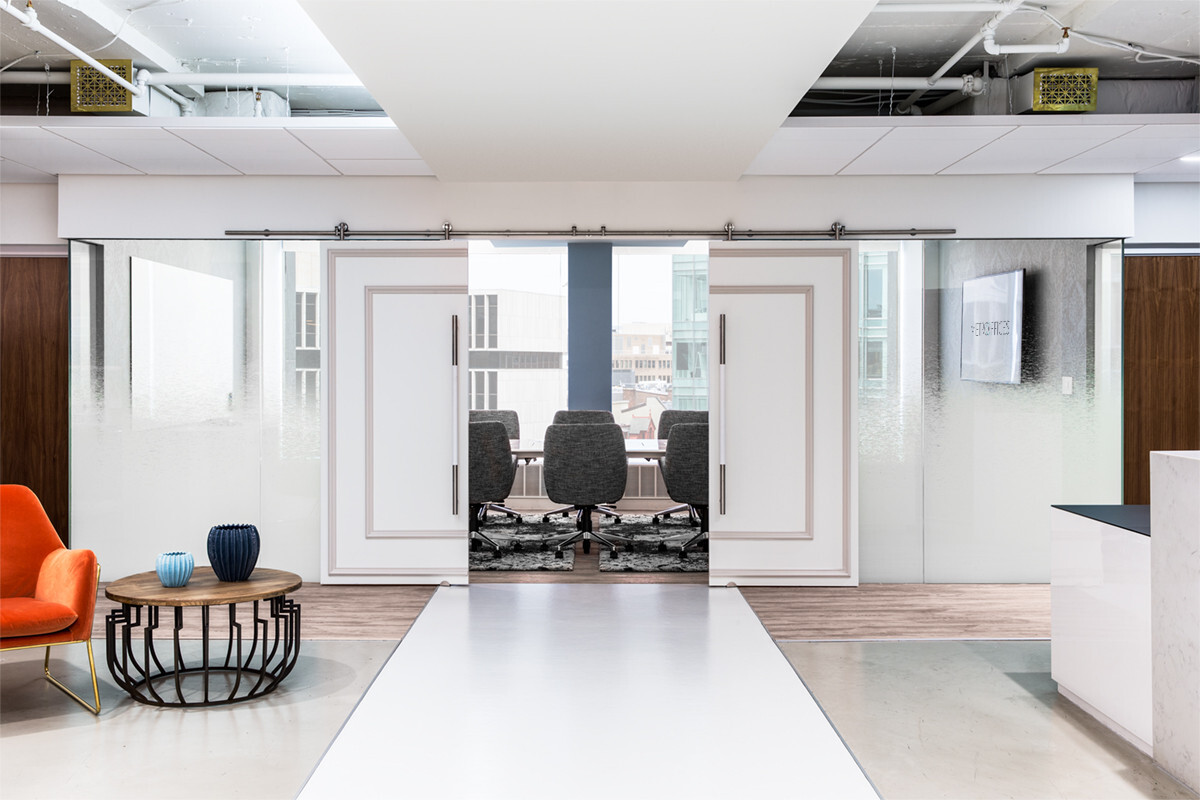 Modern office space with a corridor leading to a glass-walled conference room furnished with gray chairs. The corridor has a white ceiling and floor, with an orange chair and side table on the left side.