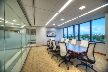 A modern conference room with a long table, surrounded by office chairs, a large window offering an exterior view, and a wall-mounted screen displaying a news channel.