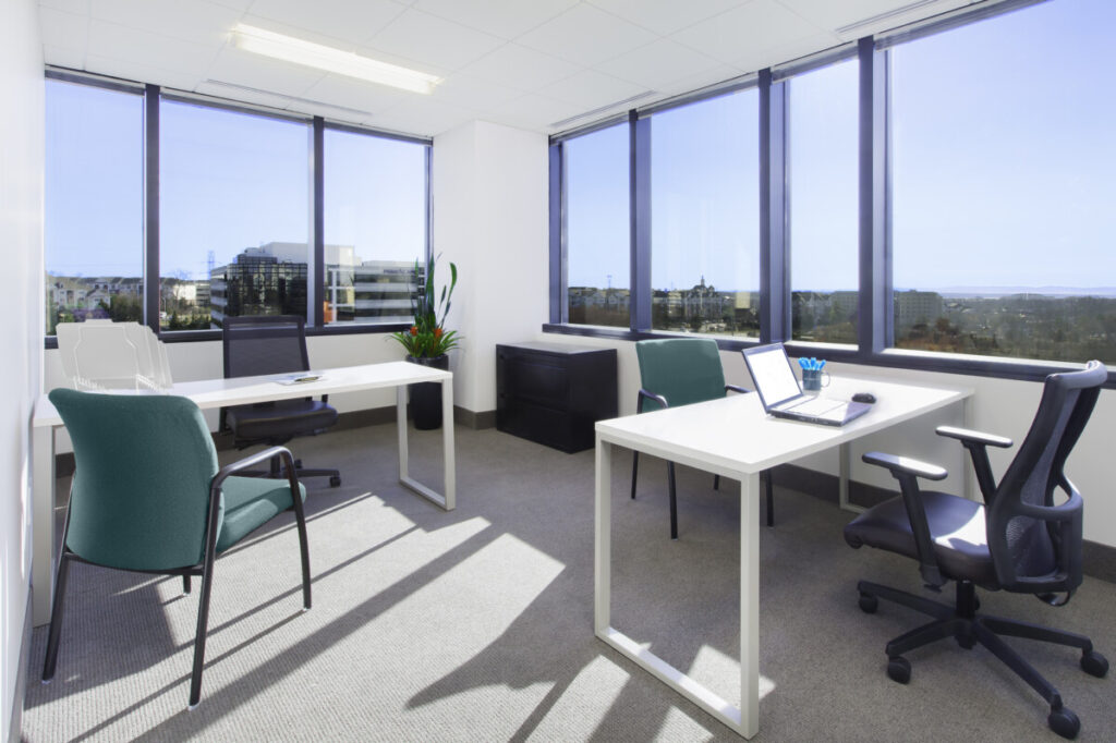 A bright office with two desks, chairs, laptops, and a potted plant. Large windows provide natural light and a view of city buildings.