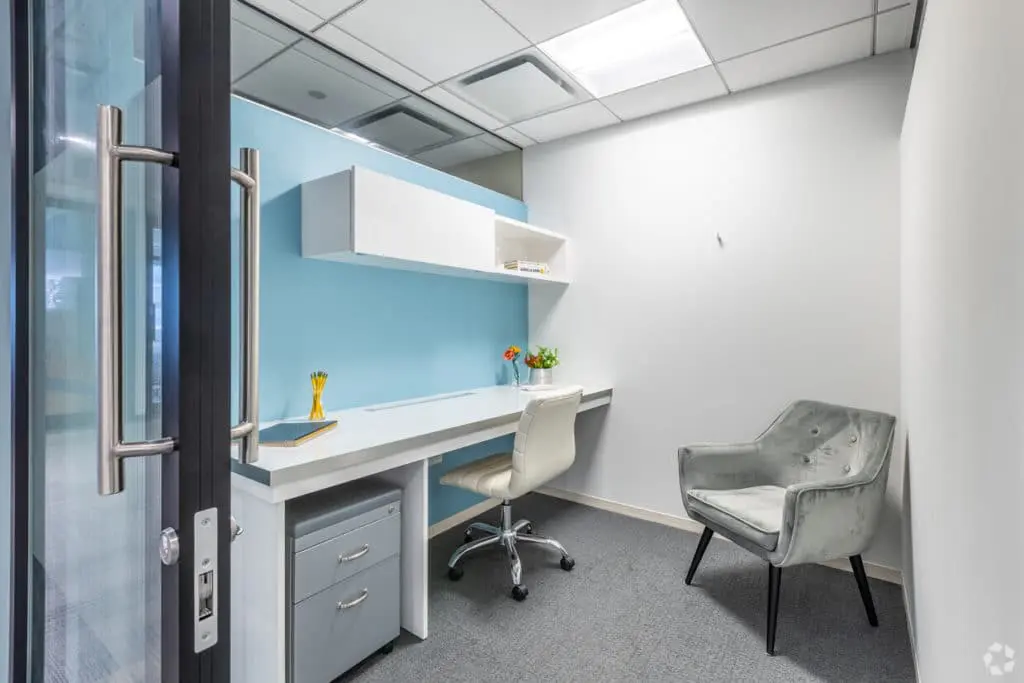 Modern office space featuring a white desk with shelves, a rolling chair, and a grey armchair, viewed through an open door.