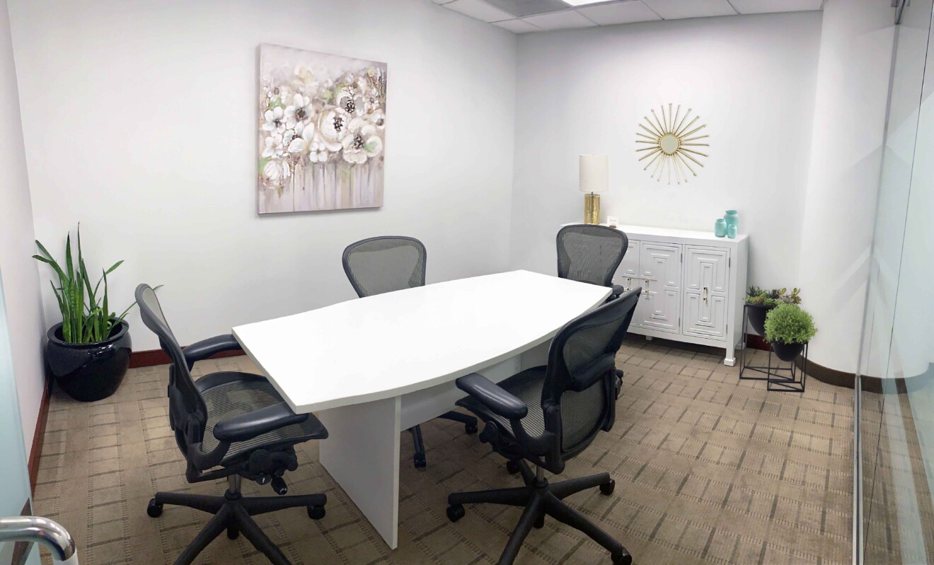 A small conference room with a white table, four black chairs, a plant, a painting on the wall, and a sideboard with decorative items. The floor is carpeted, and the room is well-lit.