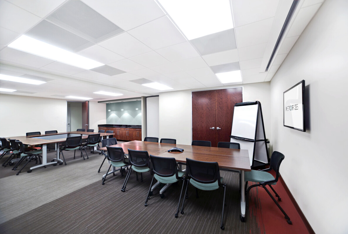 A modern conference room with multiple tables, chairs, a presentation screen, a whiteboard, and a closed door in the background.