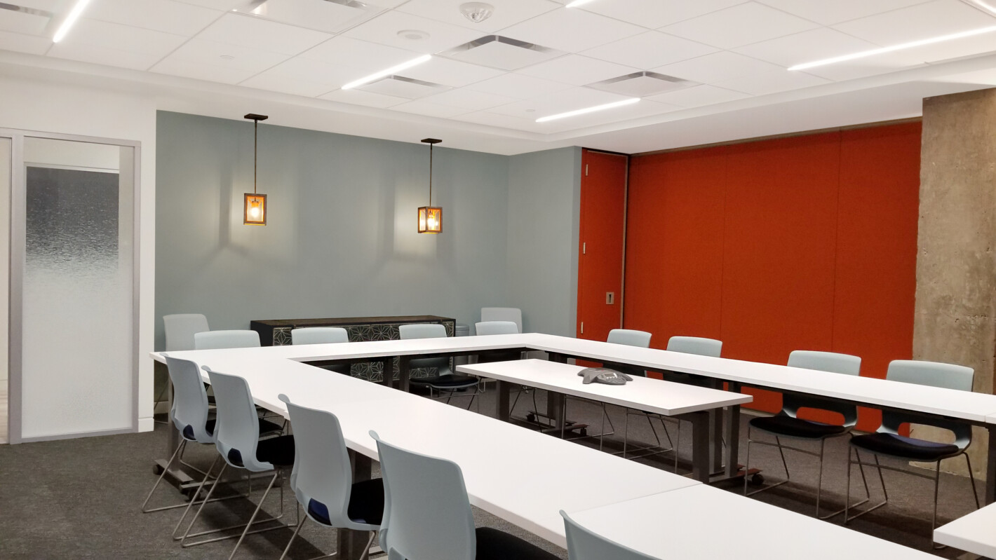 A modern conference room with U-shaped white tables, light blue chairs, red accent wall, and frosted glass door. Two ceiling lights illuminate the room.