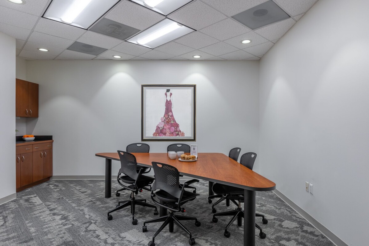 A conference room with a long wooden table, five black swivel chairs, a framed painting on the wall, and cabinets in the corner. Bowl of fruit and informational stand on the table.