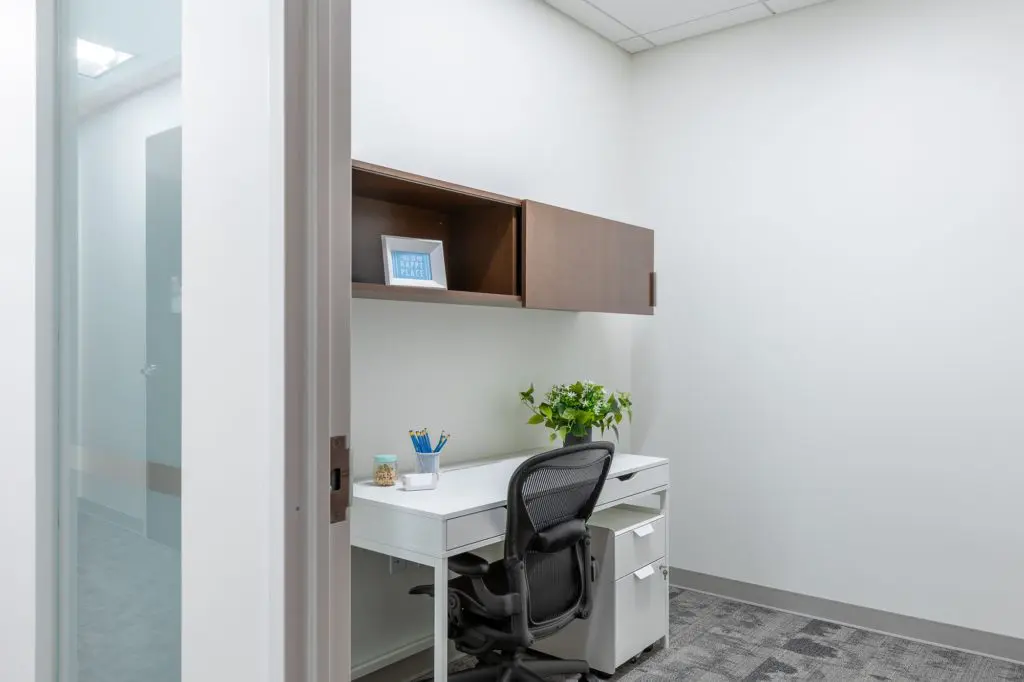 A modern office space featuring a white desk with a black office chair, wooden cabinets, a potted plant, and decor items, viewed through a glass door.