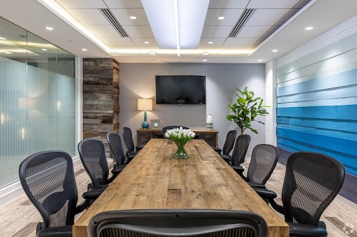 A modern conference room with a long wooden table, eight black mesh chairs, a wall-mounted screen, a plant, and a lamp. The walls feature glass panels and blue accents.