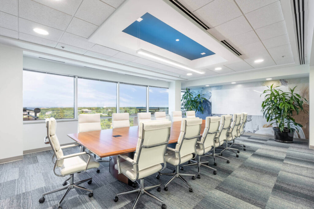 A modern conference room with a long wooden table, surrounded by white office chairs, large windows providing natural light, and a ceiling with a blue accent.