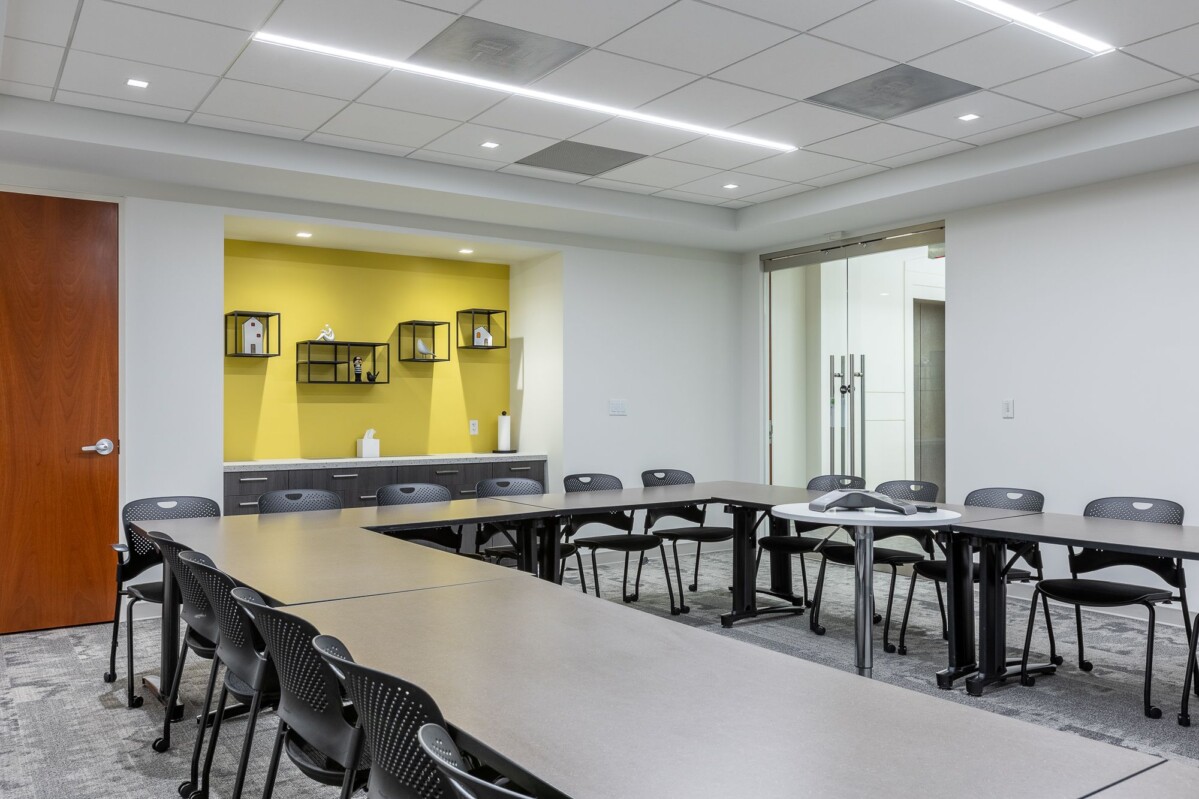 The Shenandoah Training Room boasts a contemporary conference environment, complete with U-shaped rectangular tables, numerous black chairs, a wooden door, and a yellow accent wall adorned with shelves.