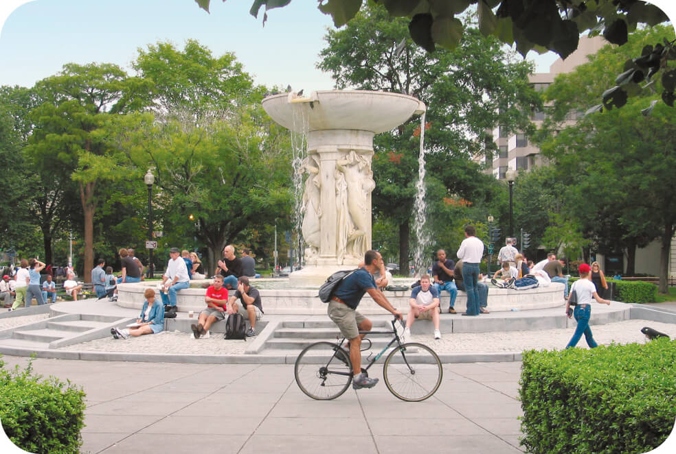 Visitors unwind as a cyclist navigates the path near a grand fountain in Central Park, embraced by lush trees and abundant greenery.