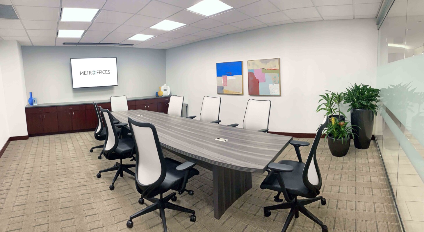 A modern conference room with a long table, eight chairs, a screen displaying 'METRO FACES,' and wall art. The room has carpeting, an indoor plant, and concealed lighting.