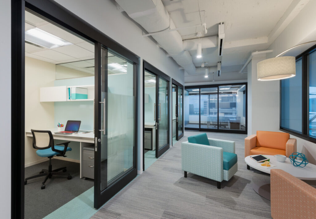 Modern office space with glass-walled private offices, ergonomic chairs, a communal seating area with armchairs, and a small table. The large windows provide ample natural light, making it ideal for those working in shared spaces.