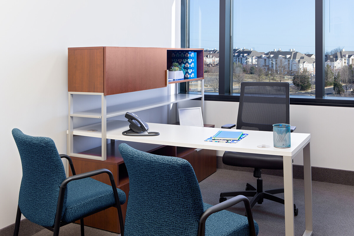 Modern office with a desk, chair, telephone, notebook, pen, and a cup, flanked by two visitor chairs. A window offers a view of a cityscape and trees. Shelves and cabinets are visible behind the desk.