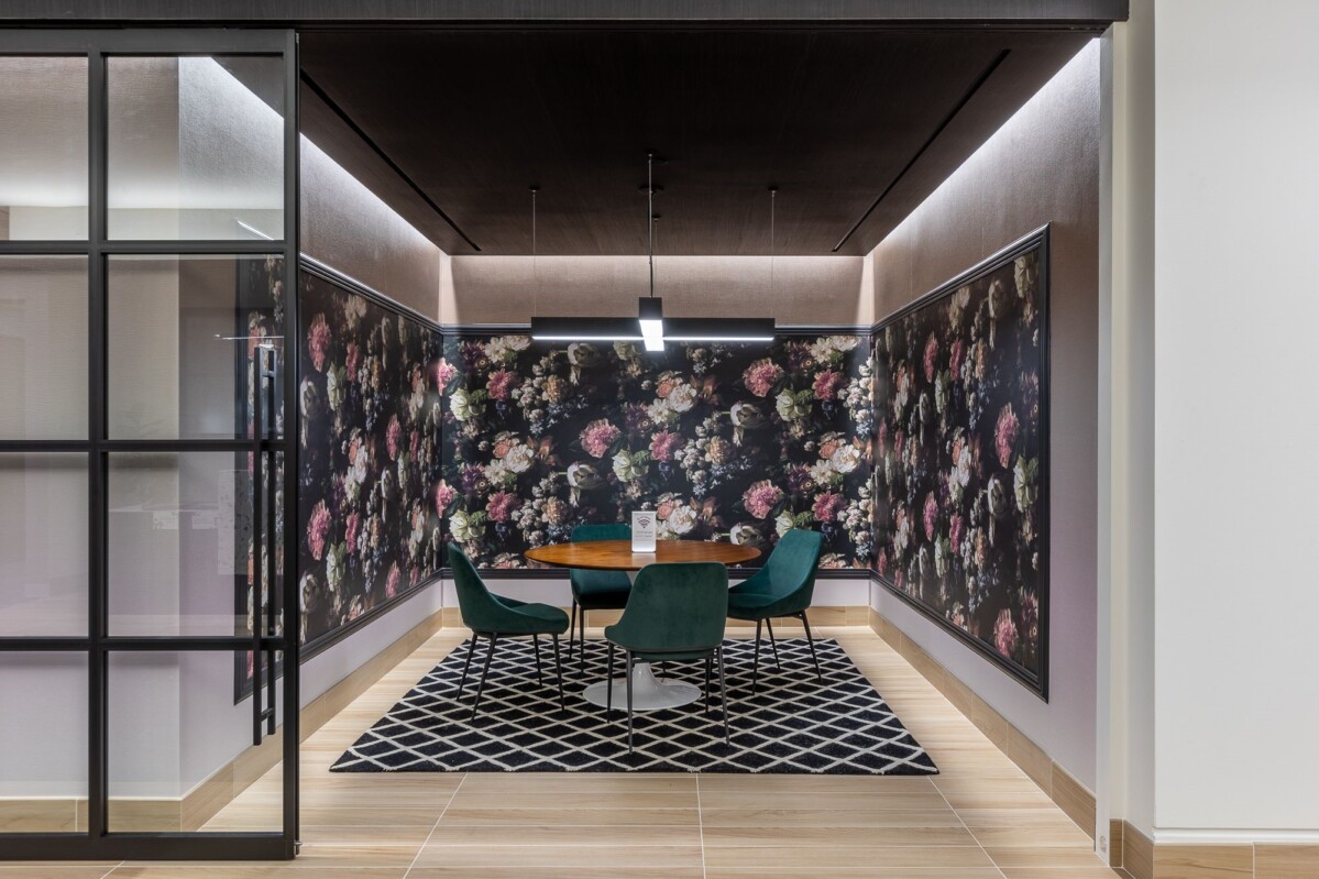 Modern meeting room with a round wooden table, four green chairs, and floral wallpaper. The room has a geometric rug and is enclosed by glass walls.