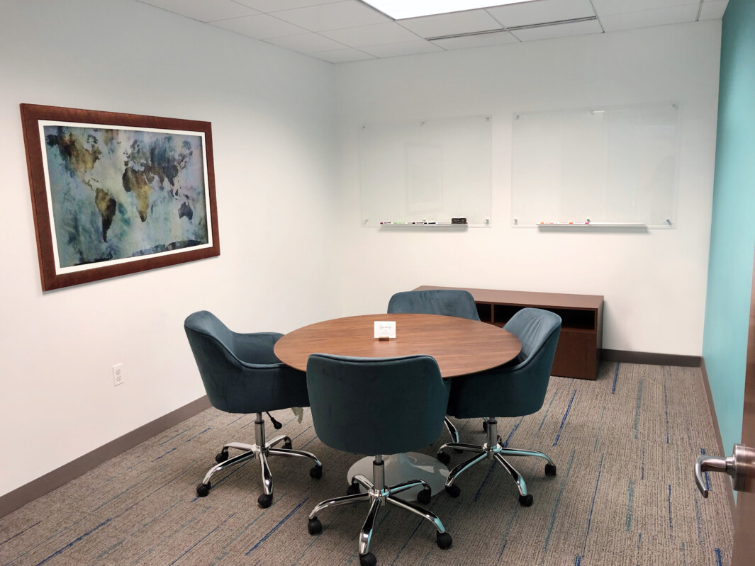 A small conference room with a round table, four chairs, a world map artwork hanging on the wall, and two whiteboards.