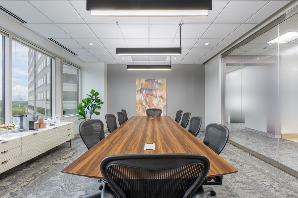 A modern conference room with a long wooden table, black chairs, large windows on the left, abstract wall art, a potted plant, a sideboard with snacks and a glass partition on the right.