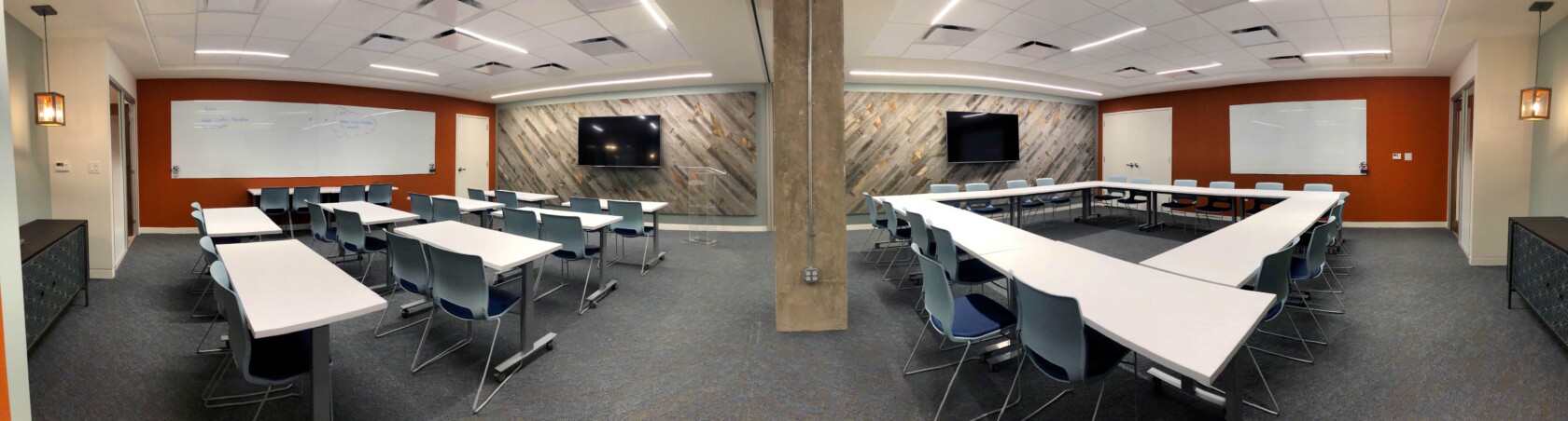 Panoramic view of a modern classroom featuring desks, chairs, whiteboards, and large monitors on the walls. The room is brightly lit with overhead lights and has a combination of red and wooden paneled walls.