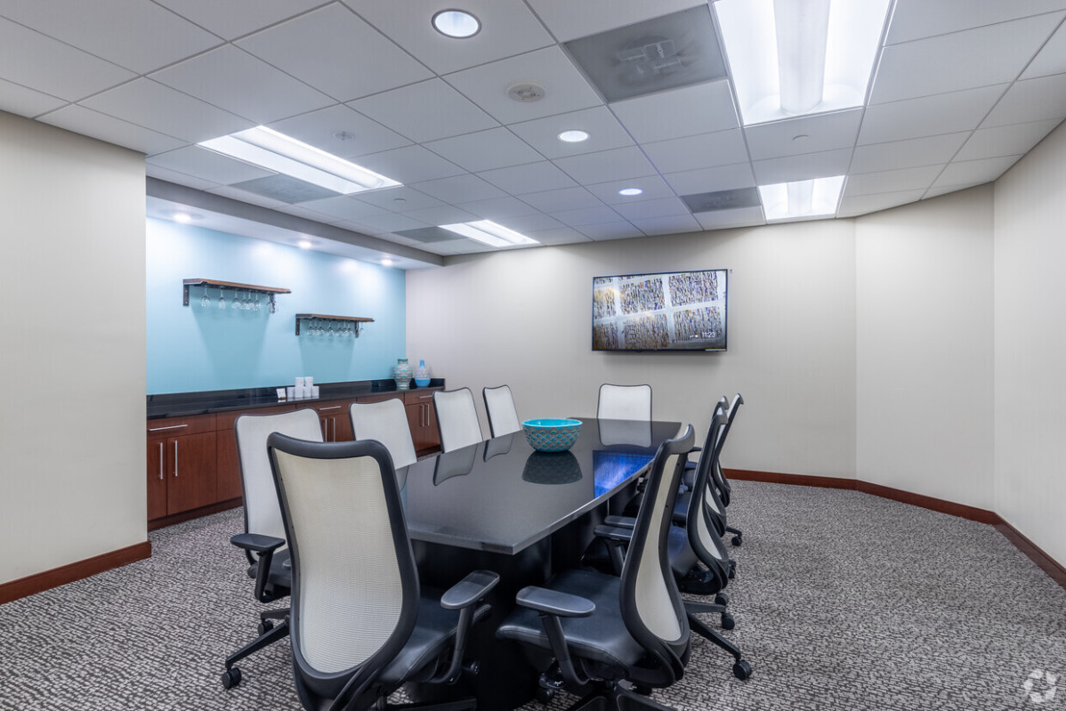 A modern conference room with a long black table, eight white office chairs, a wall-mounted TV, shelves, and a blue decorative bowl on the table. The room has overhead lighting and carpeted flooring.