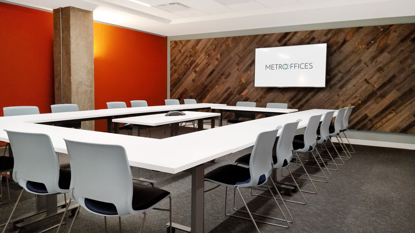 A modern conference room with a large U-shaped table, several gray chairs, a TV on the wall displaying "Metro Offices," and orange and wooden decor.