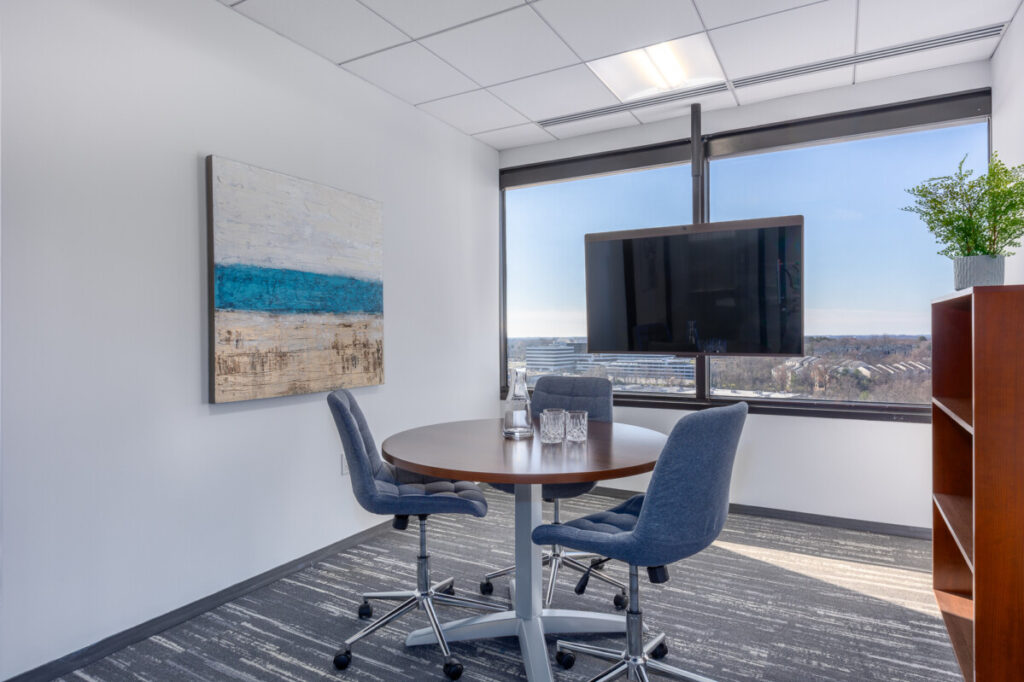 A small office with a round table, three chairs, a mounted TV, a framed painting on the wall, and a bookshelf with a potted plant. It's the perfect setup for those used to working in shared environments, enhanced by a window offering an impressive city view.